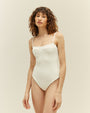 An angled knee-up view of a brunette model wearing a white one-piece swimsuit with a seam at the upper chest and thin straps, standing in front of a white background.