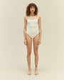 A full body view of a brunette model wearing a white one-piece swimsuit with a seam at the upper chest and thin straps, standing in front of a white background.