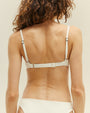 A waist-up backside view of a brunette model with her hair pushed to the side, wearing a white bikini with thin straps and a horizontal line in the middle of the top. The background is plain white.