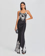 A full body view of a brunette model wearing a thin strapped round neck black dress with a white marbled design at the top and bottom of the dress. She is accessorizing with a silver clutch and round silver earrings. The background is plain white.