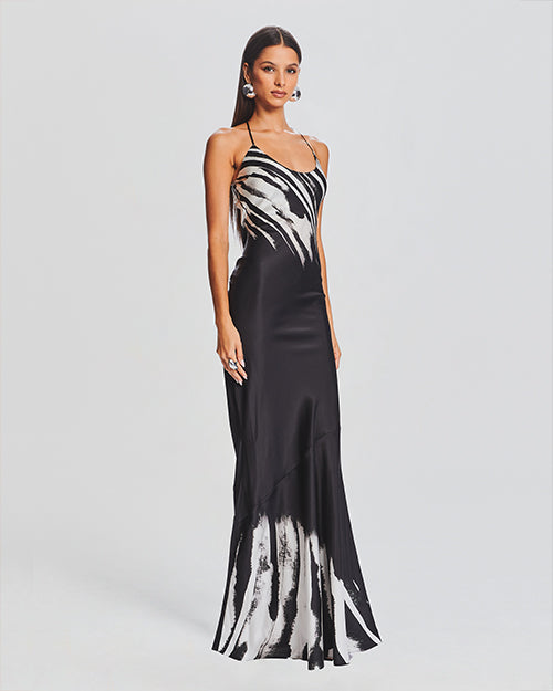 A full body sideview of a brunette model wearing a thin strapped round neck black dress with a white marbled design at the top and bottom of the dress. She is accessorizing with a silver clutch and round silver earrings. The background is plain white.