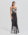 A full body sideview of a brunette model wearing a thin strapped round neck black dress with a white marbled design at the top and bottom of the dress. She is accessorizing with a silver clutch and round silver earrings. The background is plain white.