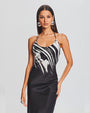 A knee-up view of a brunette model with her hand on her hip wearing a thin strapped round neck black dress with a white marbled design at the top, a silver ring and round silver earrings. The background is plain white.