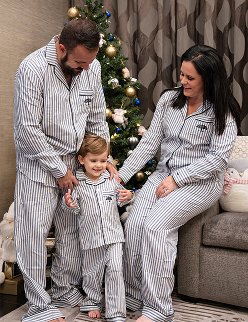 A barefoot woman, man and a child in a living room setting with a Christmas tree and presents behind them. Everyone is wearing matching blue and white striped pajamas with a blue tree logo on the chest pocket.