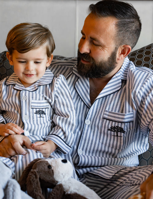 A little boy with a dog toy sitting in a man's lap, both wearing matching blue and white striped button-down pajamas with a blue tree logo on the chest pocket. They are sitting in front of multiple pillows and a white wall.