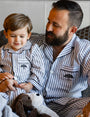 A little boy with a dog toy sitting in a man's lap, both wearing matching blue and white striped button-down pajamas with a blue tree logo on the chest pocket. They are sitting in front of multiple pillows and a white wall.
