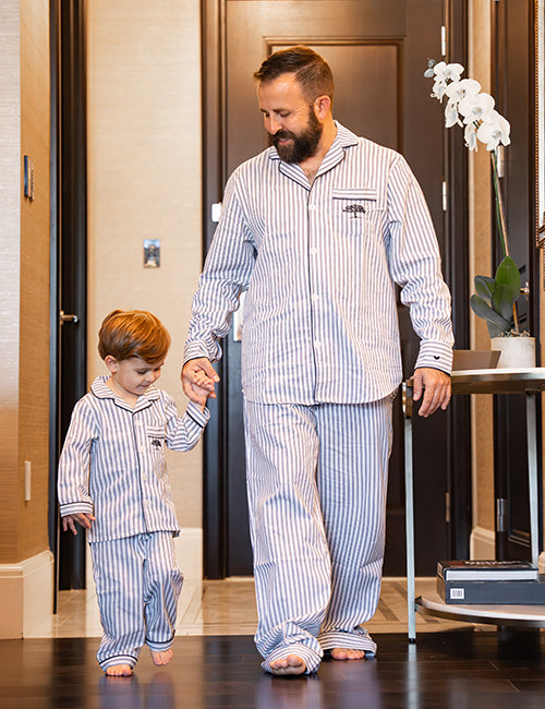 A barefoot man and a child walking hand in hand inside of a home setting, both wearing matching blue and white striped button-down pajamas with long-sleeves and a blue tree logo on the chest pocket.
