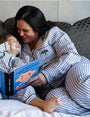 A little boy and his mom sitting on a bed reading a book called "PABLO PICASSO". Both models are wearing matching blue and white striped button-down pajamas with a blue tree logo on the chest pocket. They are sitting in front of multiple pillows and a white wall.