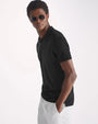 A torso shot of a model with his hand in his pocket, standing against a white background and wearing a black short-sleeve polo shirt with square sunglasses and white pants.