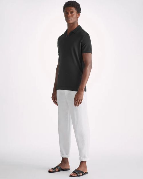 A full body shot of a model standing against a white background, wearing a black short-sleeve polo shirt with white pants. The model is also wearing white rolled up pants and black open-toed sandals.