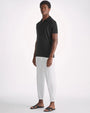 A full body shot of a model standing against a white background, wearing a black short-sleeve polo shirt with white pants. The model is also wearing white rolled up pants and black open-toed sandals.