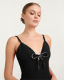 A closer waist-up view of a model wearing a long black gown with thin shoulder straps, and a deep V-neckline adorned with a decorative bow at the center. The background is white.