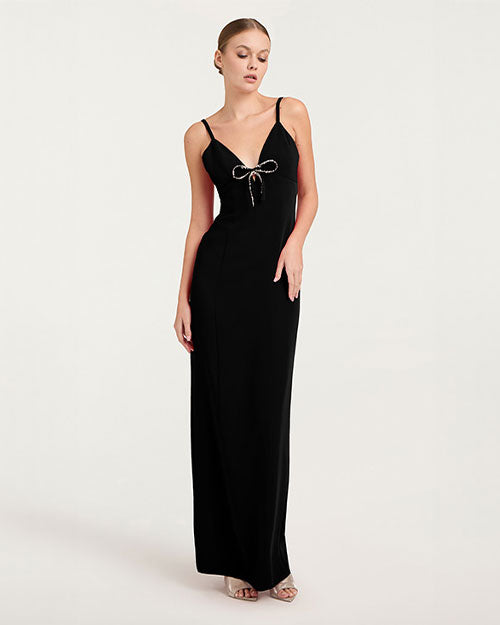 A full body shot of a model wearing a long black gown with thin shoulder straps, and a deep V-neckline adorned with a decorative bow at the center. The model is wearing beige open-toed heels and standing in front of a white background.