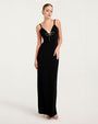 A full body shot of a model wearing a long black gown with thin shoulder straps, and a deep V-neckline adorned with a decorative bow at the center. The model is wearing beige open-toed heels and standing in front of a white background.