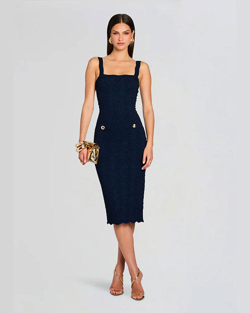 A stylish woman in a fitted navy dress with square straps and gold button accents, holding a gold clutch and wearing open-toe heels.