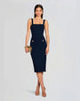 A stylish woman in a fitted navy dress with square straps and gold button accents, holding a gold clutch and wearing open-toe heels.