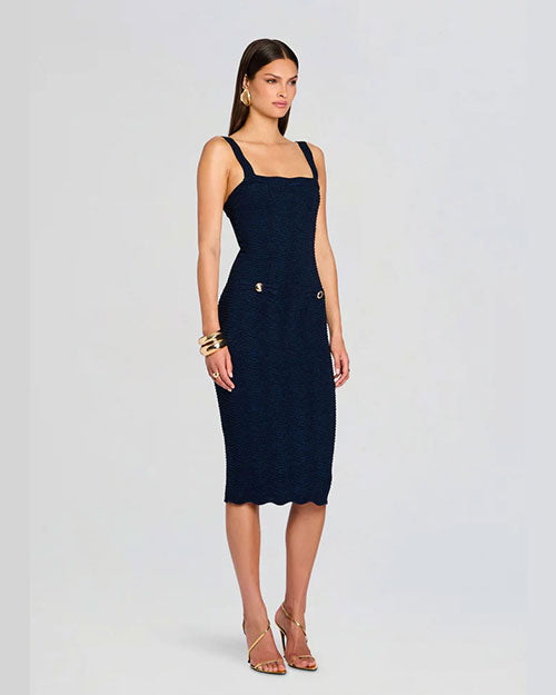 A woman poses in a three-quarter stance wearing a fitted navy dress with a square neckline, gold accents, and strappy heels. She faces forward with her full face visible, set against a light background.
