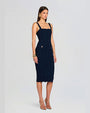 A woman poses in a three-quarter stance wearing a fitted navy dress with a square neckline, gold accents, and strappy heels. She faces forward with her full face visible, set against a light background.