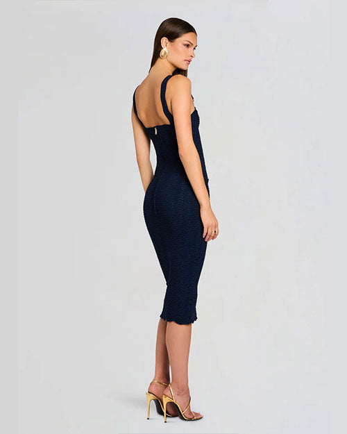 A woman in a fitted navy dress and strappy heels poses in a three-quarter turn, showing mostly the back of the dress and her face in profile. She is not looking at the camera and stands against a neutral background, highlighting the dress's elegant silhouette.