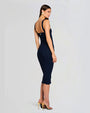 A woman in a fitted navy dress and strappy heels poses in a three-quarter turn, showing mostly the back of the dress and her face in profile. She is not looking at the camera and stands against a neutral background, highlighting the dress's elegant silhouette.