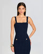Close-up of a brunette woman modeling a fitted navy dress with a square neckline and gold button accents, set against a grey background. Her legs are not visible in the frame, emphasizing the dress and elegant accessories.
