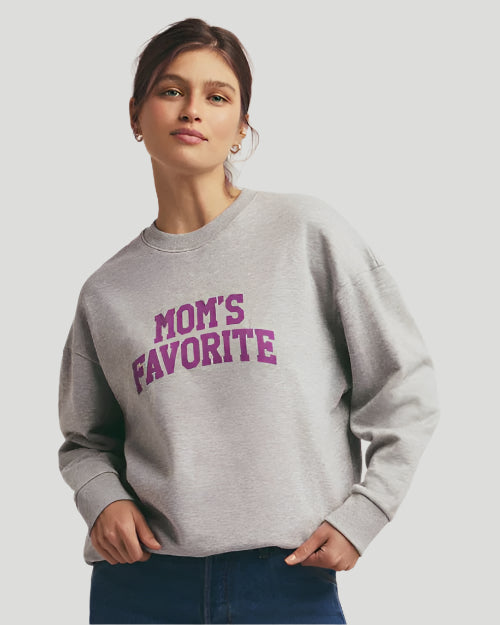 A waist-up view of a brunette model wearing a light gray crewneck sweatshirt with the text "MOM'S FAVORITE" printed in bold. The model paired the sweatshirt with blue jeans and gold hoops and is standing in front of a grey background.