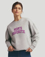 A waist-up view of a brunette model wearing a light gray crewneck sweatshirt with the text "MOM'S FAVORITE" printed in bold. The model paired the sweatshirt with blue jeans and gold hoops and is standing in front of a grey background.