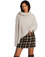 A knee-up view of a dark-haired model wearing a light-colored, long-sleeved sweater with a matching scarf, and a brown plaid skirt with knee-high black boots. The background is plain white.