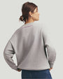 A waist-up backside view of a brunette model in a bun wearing a light gray crewneck sweatshirt with the text "FAVORITE DAUGHTER" printed in cursive under the neckline. The model is also wearing blue jeans and gold hoop earrings. The background is plain grey.