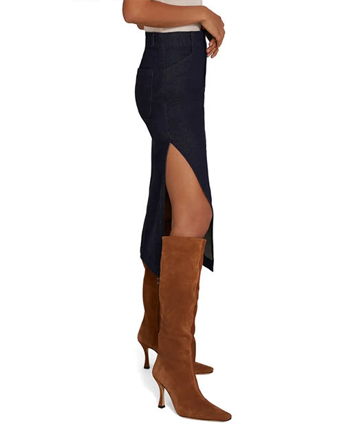 A lower body shot of a model in front of a white background wearing a long dark blue denim skirt with a high slit on the side, paired with brown suede knee-high boots.