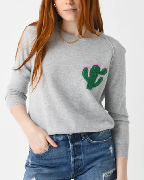 A torso shot of a ginger model wearing a light gray long-sleeve crewneck sweater with an embroidered green cactus and pink flowers on the left chest. The model is also wearing distressed blue jeans, and a gold chain with her hand in her pocket, standing in front of a white background.