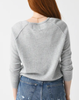 A torso level backside view of a ginger model with her hair swept to the side wearing a light gray long-sleeve crewneck sweater. The model is also wearing distressed blue jeans and a gold chain while standing against a white background.