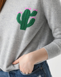A close-up image of a models' torso wearing a light gray long-sleeve crewneck sweater with an embroidered green cactus and flowers on the left chest. The model is also wearing blue jeans with her hand in her pocket.