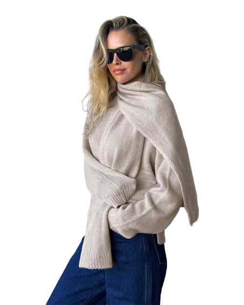 A knee-up angled view of a  model in black sunglasses wearing a beige sweater with a matching scarf and blue jeans on a white background
