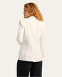 The backside view of a curly-haired model wearing a white blazer with black wide-leg pants against a white background.