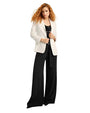 A full body shot of a curly-haired model holding the lapels of a white blazer over a black top and black wide-leg pants, held up by a flowy black belt. The model is standing against a white background.