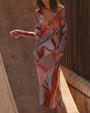 A brunette model standing in front of wooden bars and a concrete wall, wearing a long-sleeve mesh dress with a vibrant abstract multicolored print, a V-neck, an open design under the chest, and a bow at the center of the chest.
