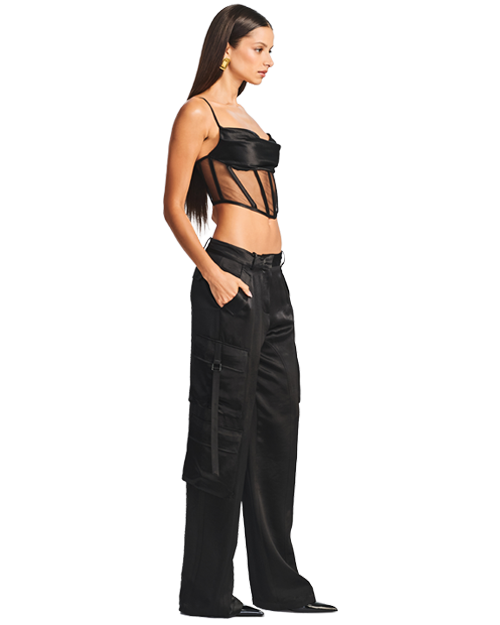 A full body sideview of a brunette model wearing a cropped, black, thin strapped sheer corset-style top with a fully black fabric chest area, paired with high-waisted black cargo pants featuring utility pockets and straps on the sides, with black pointed toe heels, and gold earrings.