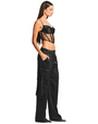 A full body sideview of a brunette model wearing a cropped, black, thin strapped sheer corset-style top with a fully black fabric chest area, paired with high-waisted black cargo pants featuring utility pockets and straps on the sides, with black pointed toe heels, and gold earrings.
