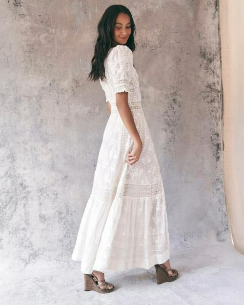 An angled, full body backside view of a dark-haired model holding wearing a long white lace dress with elbow-length ruffled sleeves with striped lace detailing, also appearing on the waistline. The model is also wearing a pair of beige open-toed heels and walking away against a grey splotchy background.