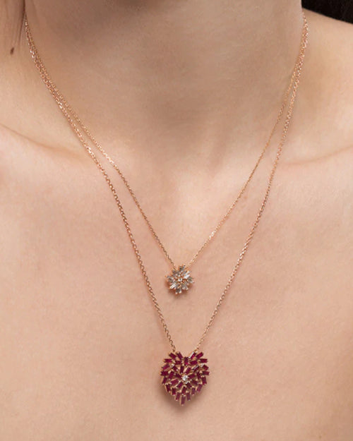 A close-up image of a models' neck wearing two gold chain necklaces, one is smaller with a crystal firework pendant, and a longer one with a pendant made out of small red rectangular diamonds, formed into a heart with a diamond in the middle.