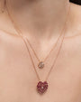 A close-up image of a models' neck wearing two gold chain necklaces, one is smaller with a crystal firework pendant, and a longer one with a pendant made out of small red rectangular diamonds, formed into a heart with a diamond in the middle.