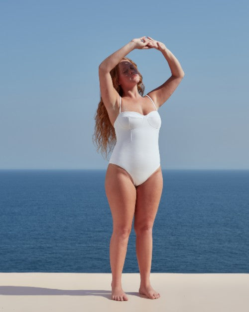 A full body view of a barefoot blonde model with her hands blocking the sun, standing in front of a blue sky and ocean, wearing a white spaghetti strapped one-piece swimsuit with a structured seam work design on the chest.