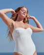 A waist-up view of a blonde model standing in front of a blue sky, with her hands touching her hair, wearing a white spaghetti strapped one-piece swimsuit with a structured seam work design on the chest.