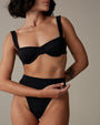 A close-up torso-level image of a short-haired model with forearm tattoos on one arm behind her back and one bare arm in front, standing in front of a beige background, wearing a black bikini top with shoulder straps and a structured seam design, including matching high-waisted bottoms.