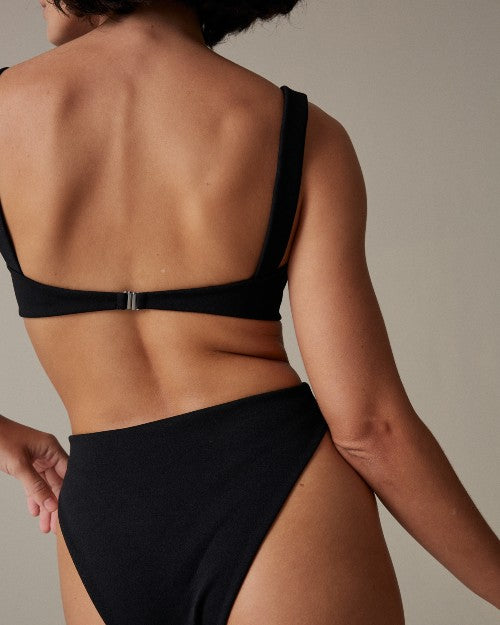 An upper body backside view of a model standing in front of a beige background, wearing a black bikini top with shoulder straps and matching high-waisted bottoms.