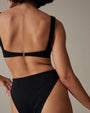 An upper body backside view of a model standing in front of a beige background, wearing a black bikini top with shoulder straps and matching high-waisted bottoms.