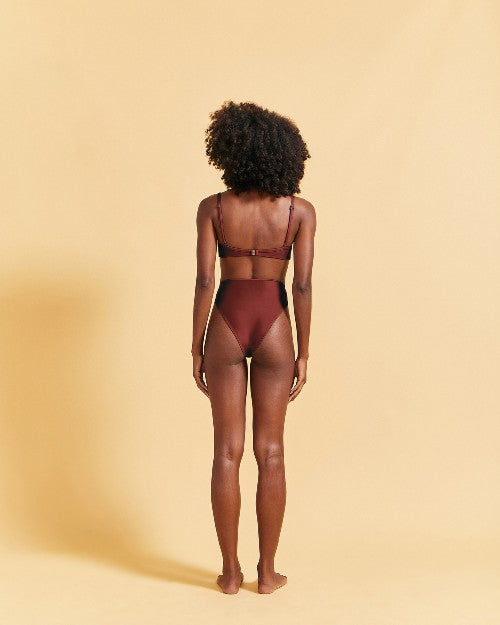 A full-body backside view of a model with her hands behind her back, wearing a brown bikini top with thin straps and matching high-waisted bottoms. The background is plain yellow.