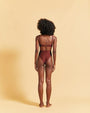 A full-body backside view of a model with her hands behind her back, wearing a brown bikini top with thin straps and matching high-waisted bottoms. The background is plain yellow.