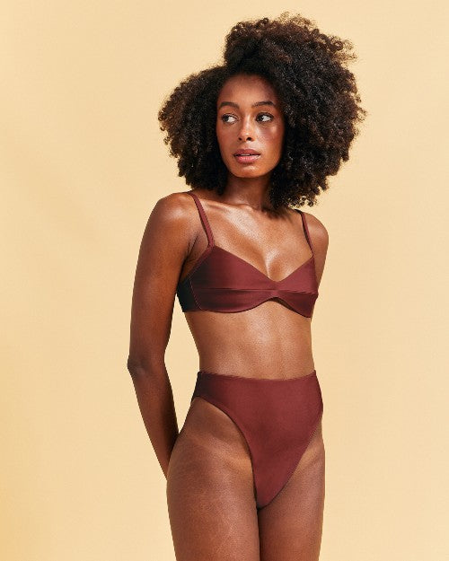 A full-body view of a model with her hands behind her back, wearing a brown bikini top with thin straps and a seam across the middle, including matching high-waisted bottoms.